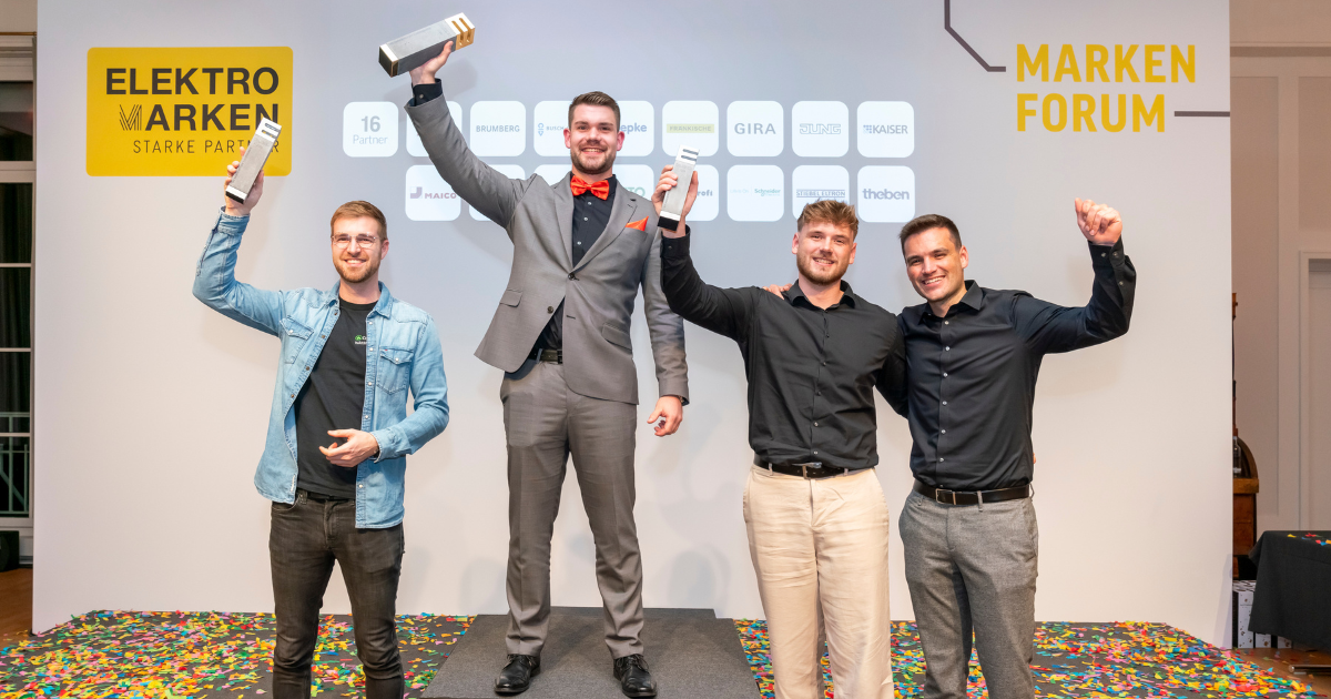 Vier junge Männer auf der Bühne des Markenforums der Elektromarken. Starke Partner. – einer steht auf dem Siegerpodest und hält stolz einen ELMAR-Award hoch, die anderen jubeln an seiner Seite. Im Hintergrund Logos der 16 Markenpartner.