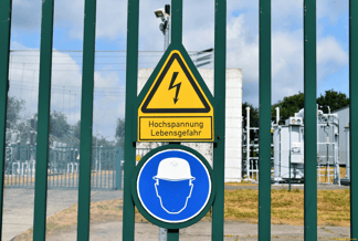 Gelbes Achtung-Schild mit Beschriftung Hochspannung Lebensgefahr auf grünem Zaun vor Trafostation