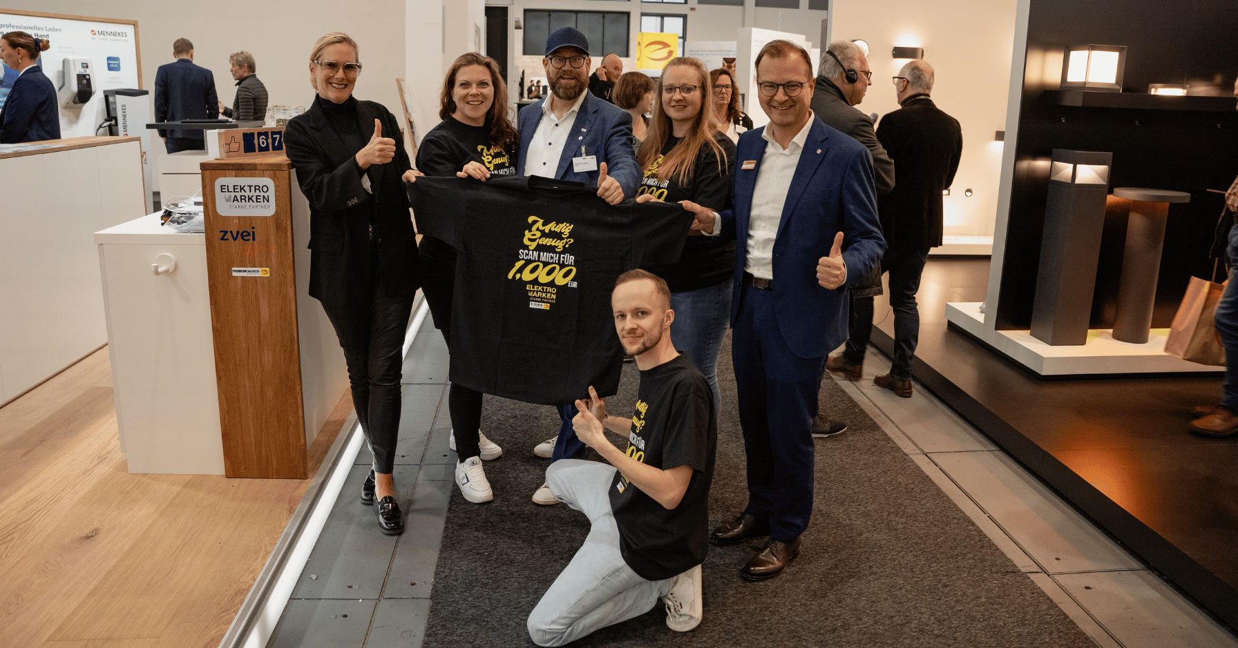 Die Creator des Elektro-Quiz Rallyes zusammen mit dem Vorstand von Elektromarken. Starke Partner auf der belektro 2024. Sie halten ein T-Shirt mit der Aufschrift 'Mutig genug? Scan mich für 1.000 EUR', das auf das Gewinnspiel der Rallye hinweist.