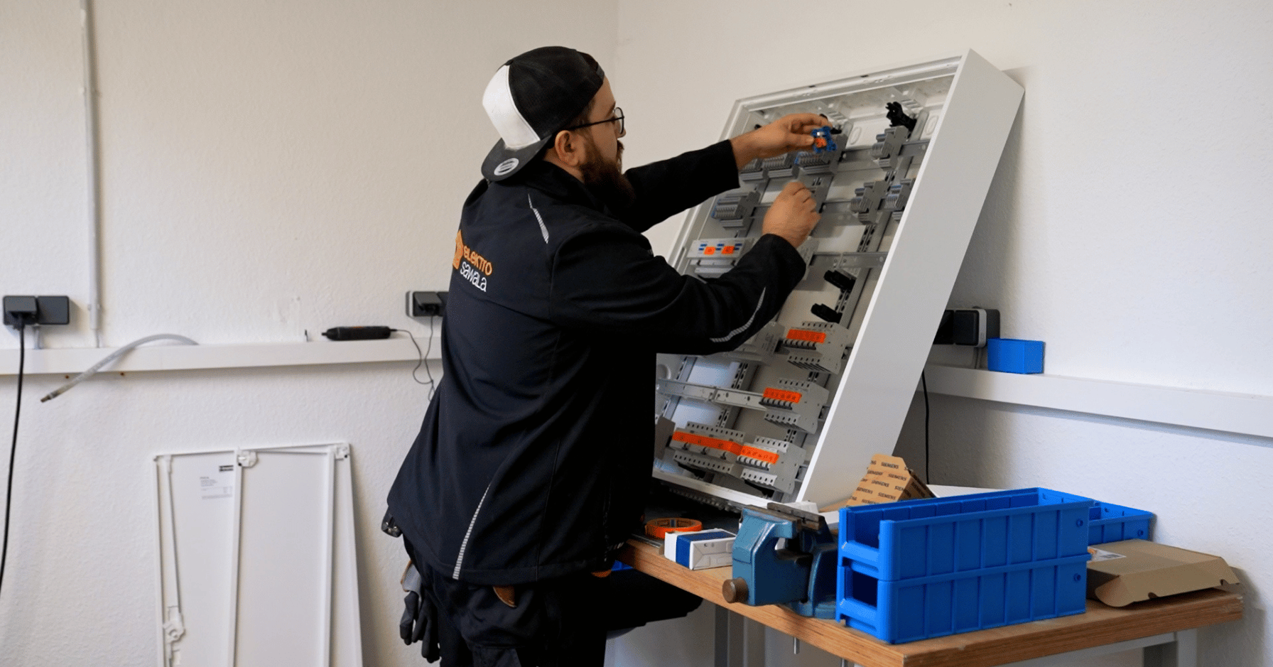 Elektriker arbeitet an einem geöffneten Schaltschrank in einer Werkstatt mit Werkzeugen und Materialboxen im Vordergrund.